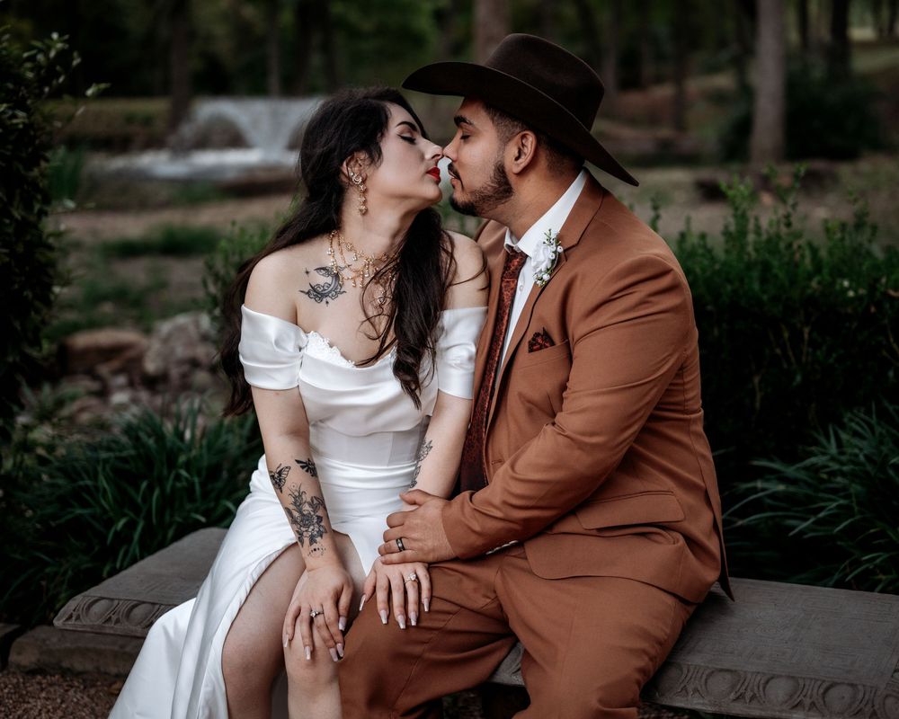 A romantic couple in wedding attire embraces near a waterfall in a lush garden setting.