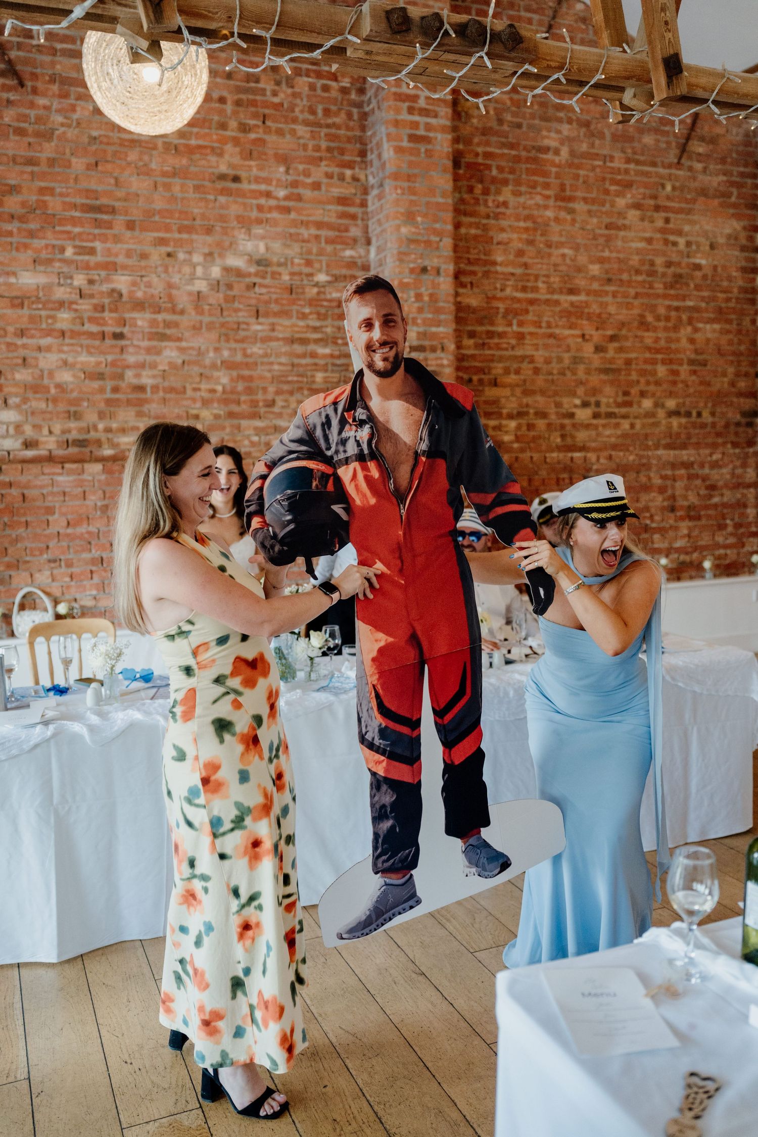 A cut out of the groom in a red jumpsuit is lifted by two women in elegant dresses at a wedding reception in a brick venue.