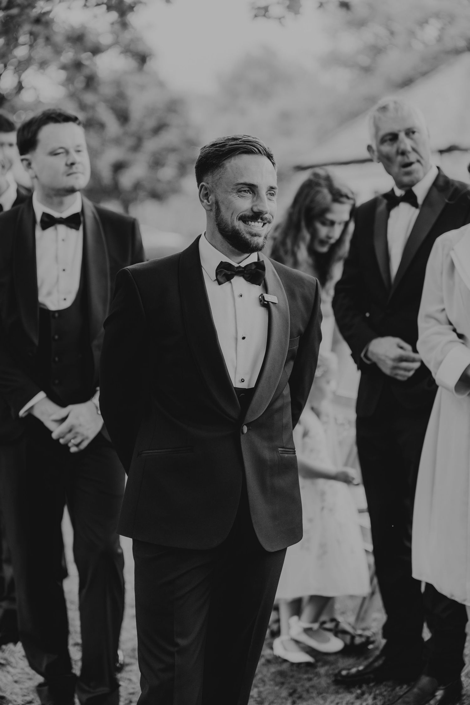 Black and white photo of a smiling groom in a tuxedo with bow tie at an outdoor wedding ceremony at The Green wedding venue in Liskeard.