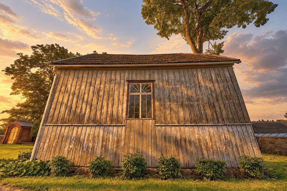 Rustic wooden barn glowing in golden sunset light with lush green shrubs and a dramatic cloudy sky backdrop.