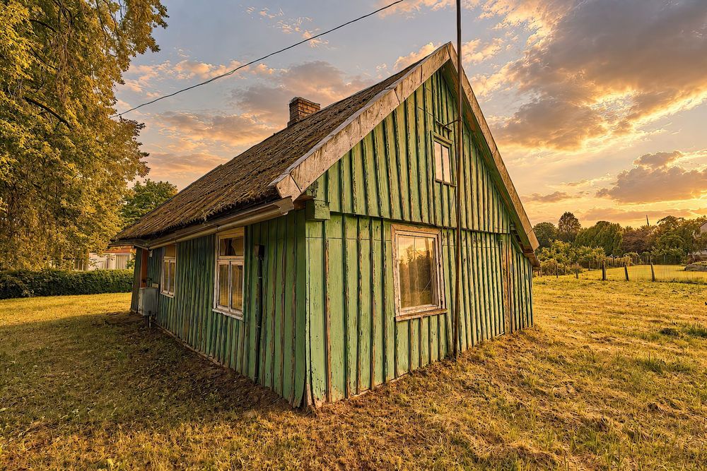 Rustic green wooden cottage with weathered siding glowing under a dramatic sunset sky surrounded by lush trees.