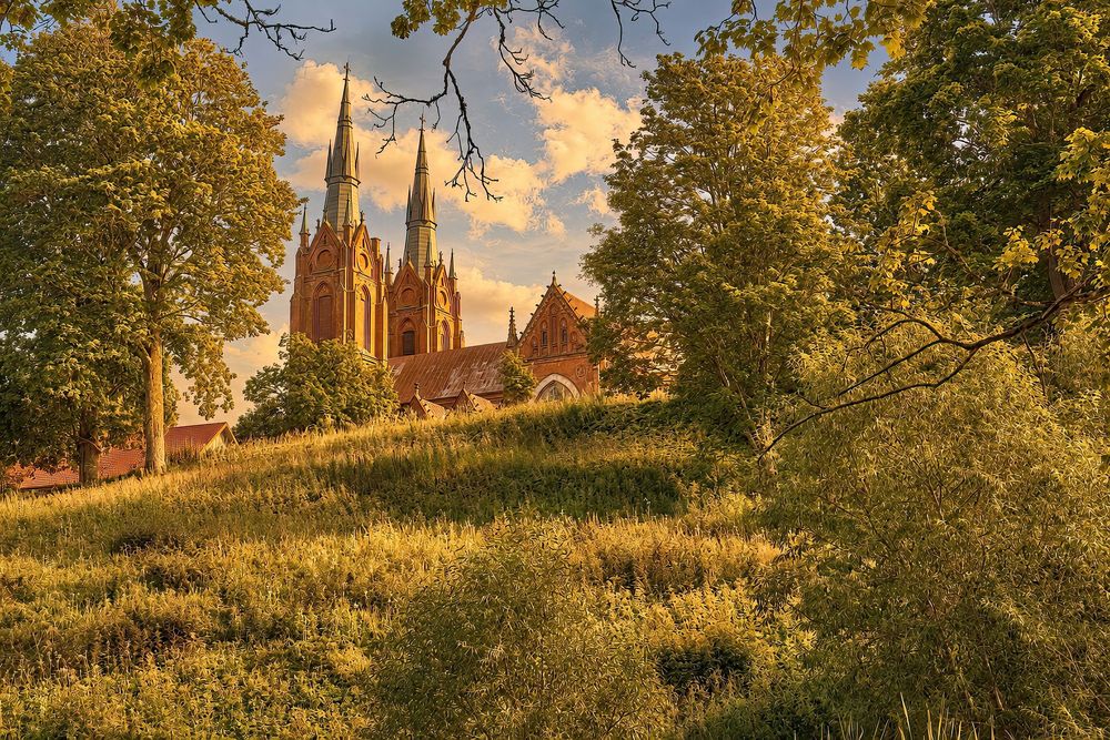 Gothic church with tall spires rises above a lush green hillside framed by trees in warm golden sunlight.