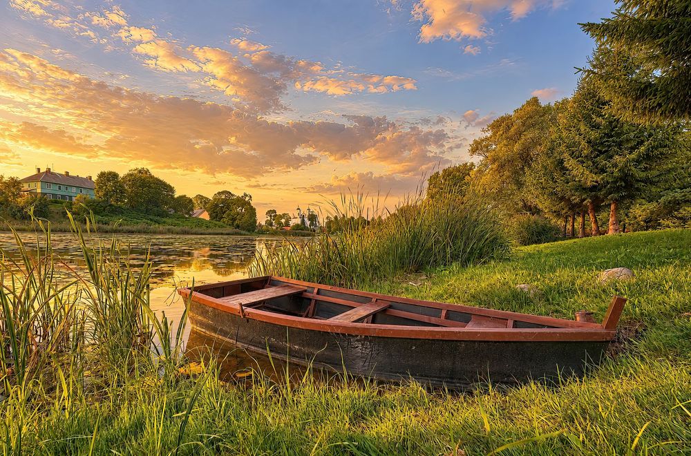 A wooden rowboat rests on a grassy riverbank at golden sunset, surrounded by reeds and lush green trees.
