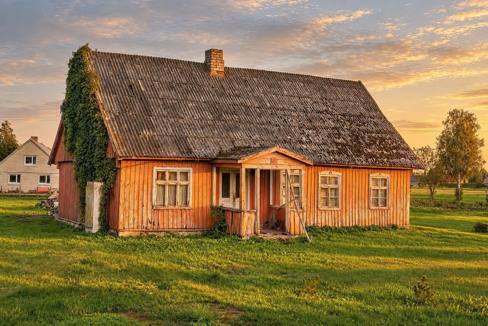 Rustic orange wooden farmhouse with ivy-covered roof at golden sunset in rural countryside setting.