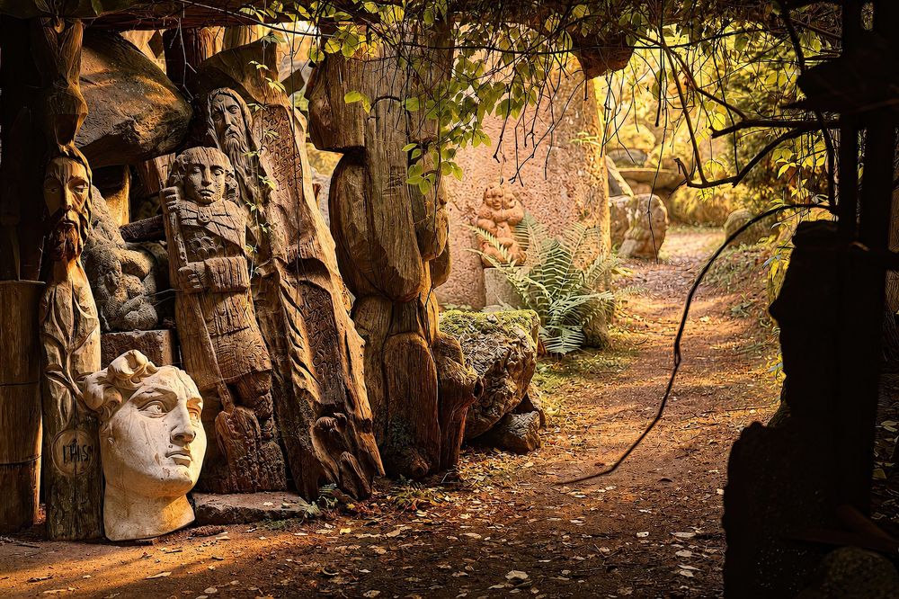 Wooden carved sculptures and stone faces line a forest path surrounded by autumn foliage and golden light.