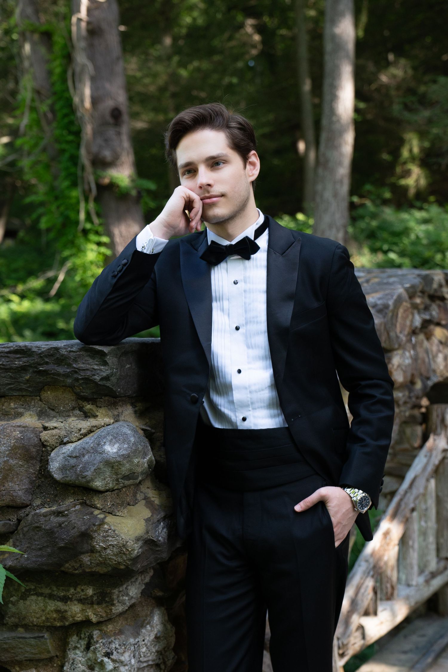 Handsome groom in black tuxedo with bow tie leaning against stone wall in forest setting.