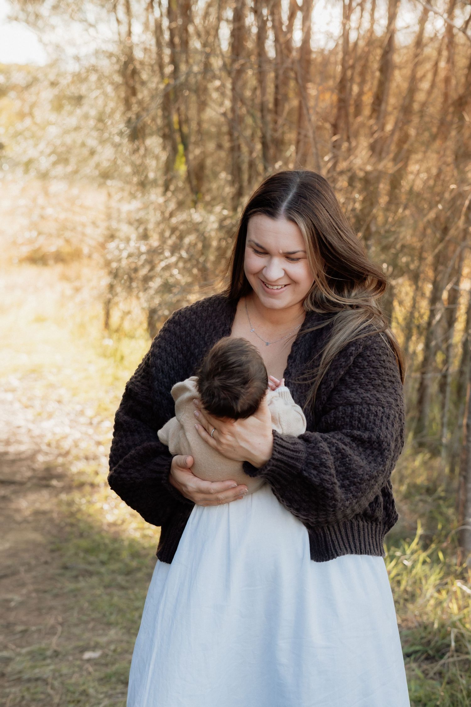 Smiling woman in dark cardigan and white skirt holds newborn baby outdoors at Rouse Hill Regional Park in golden sunlight near tree archway.