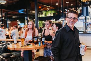 Young man smiling at bar holding beer can with group of friends socializing at high-top table in lively restaurant.