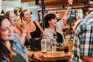 Group of women laughing and celebrating at a bar with drinks on the table in a lively restaurant setting.