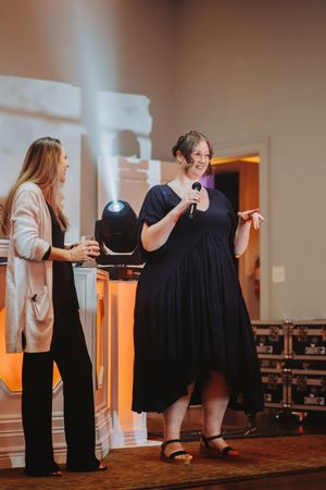 Two women on stage, one in black dress holding microphone, speaking at an event with orange ambient lighting.