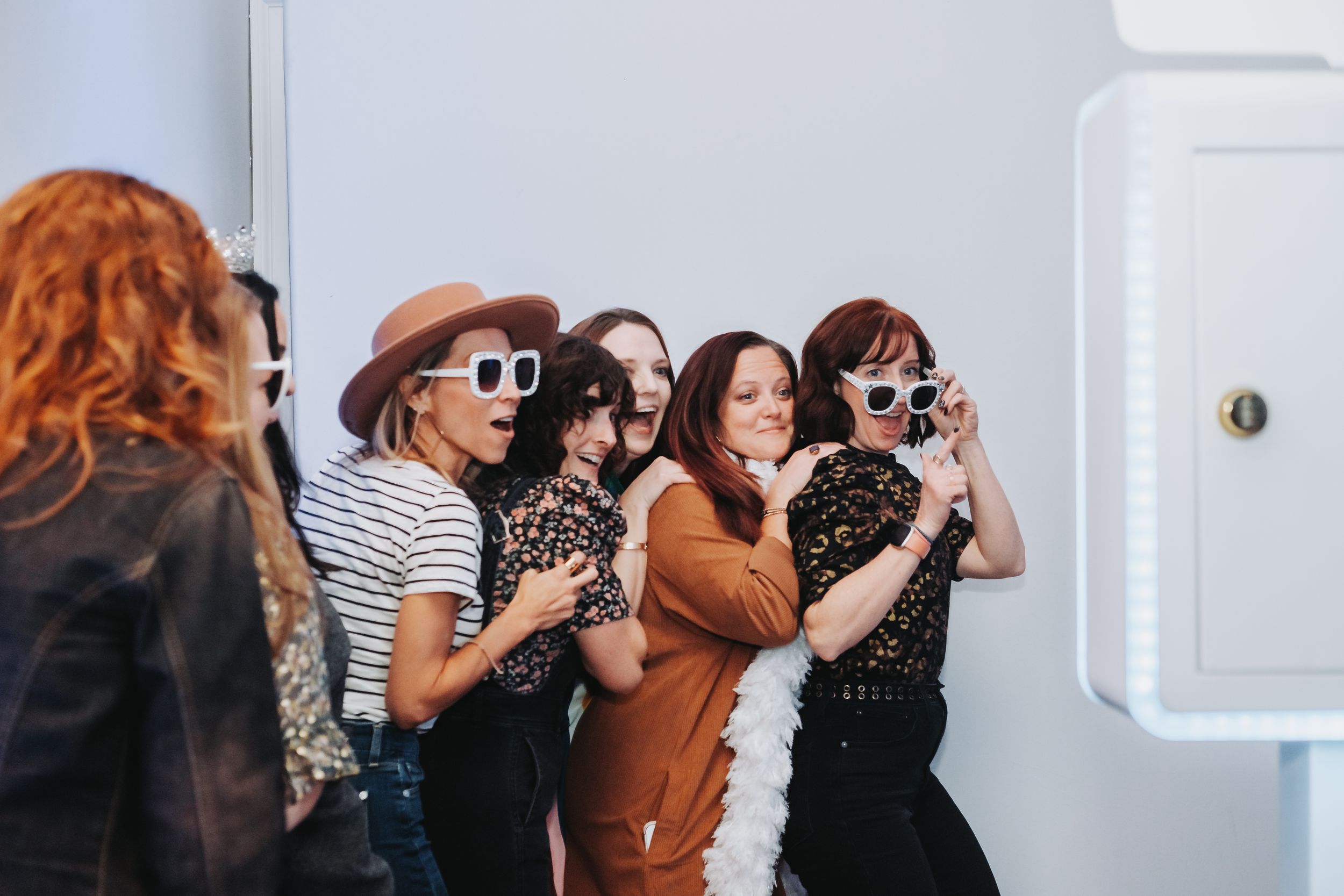 Group of women posing in a photo booth wearing sunglasses and hats, smiling and having fun at a party.