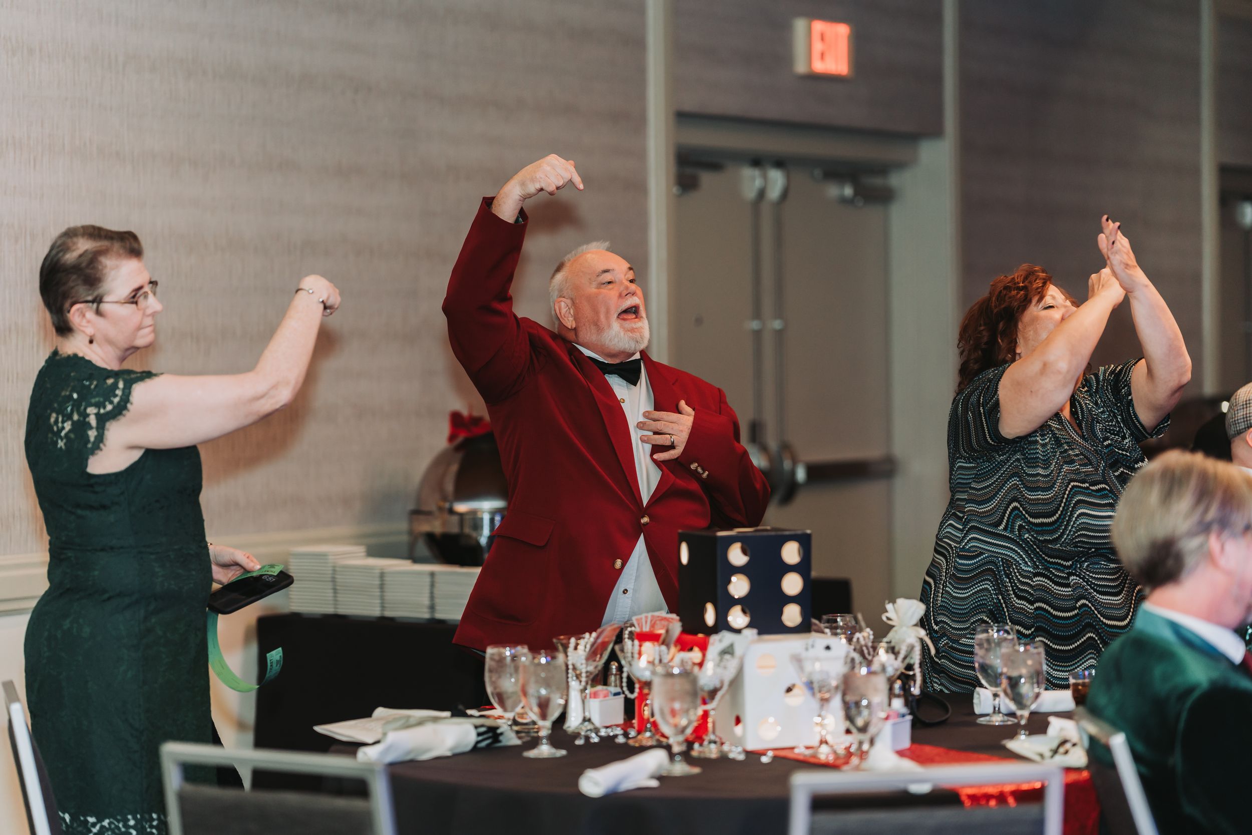 Three guests dancing joyfully at a wedding reception, man in red blazer raises arms celebratorily.