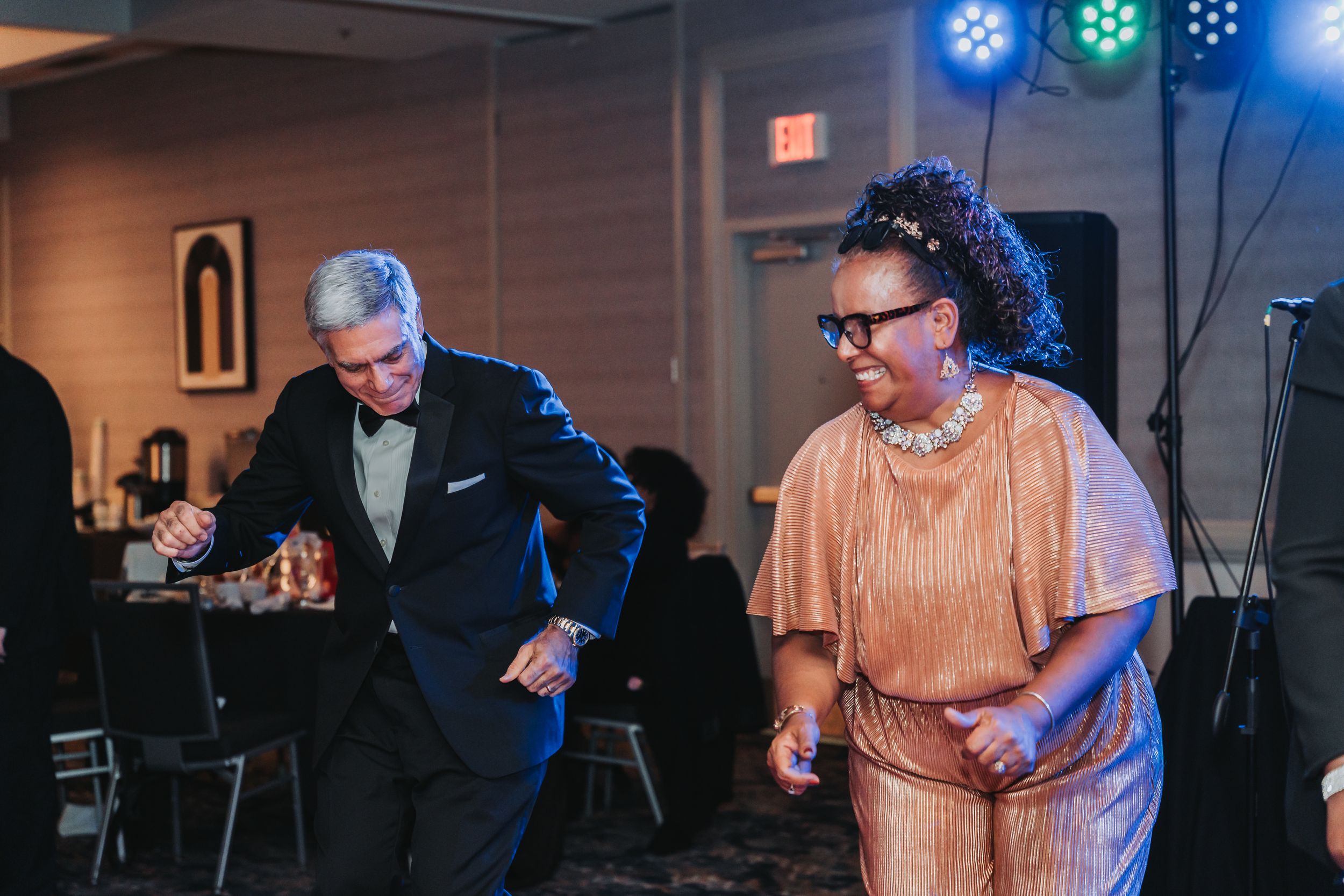 Two elegantly dressed guests dancing joyfully at a formal evening event with colorful stage lighting.