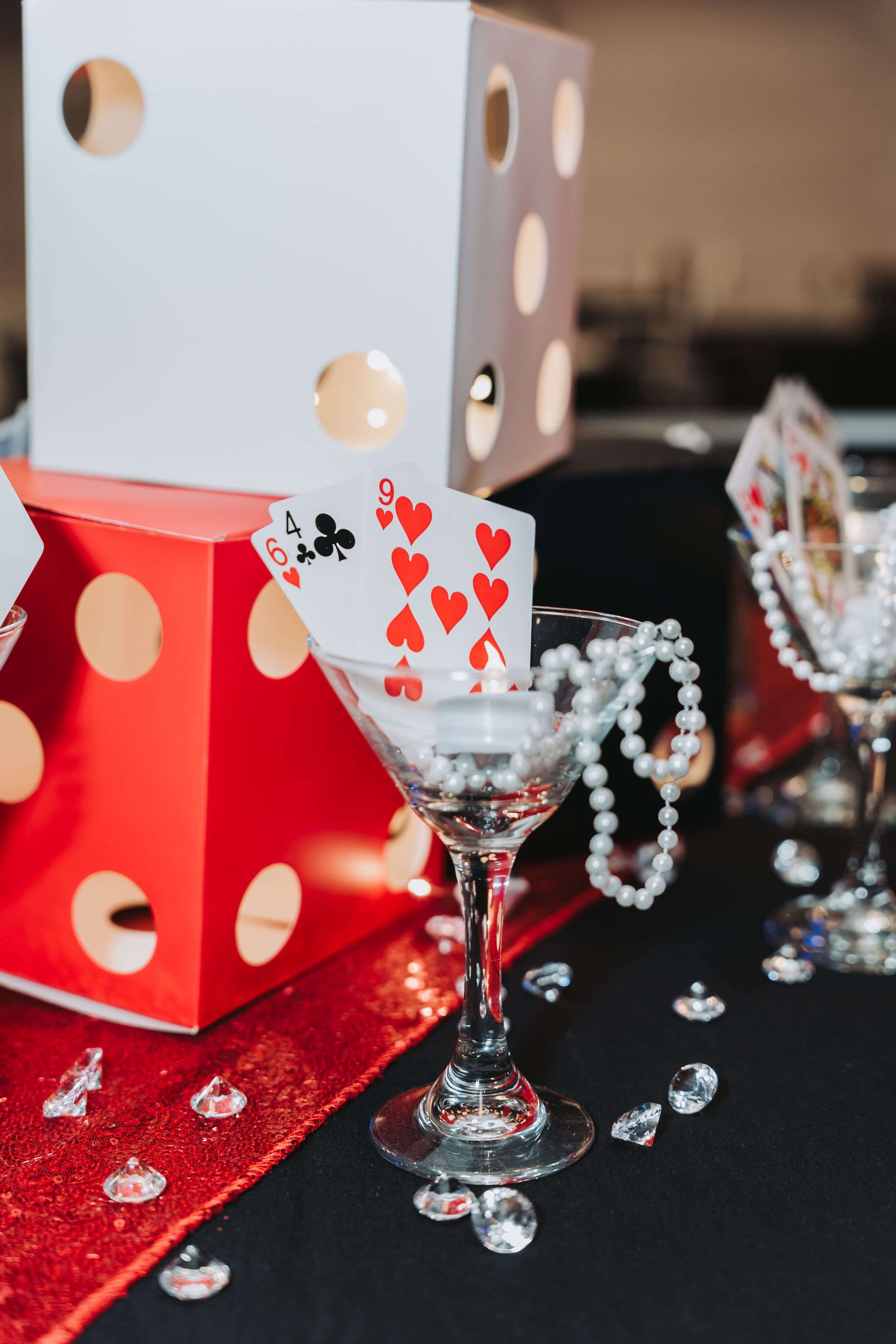 Casino-themed party decor with playing cards in martini glass, pearl beads, dice, and scattered crystals on red tablecloth.