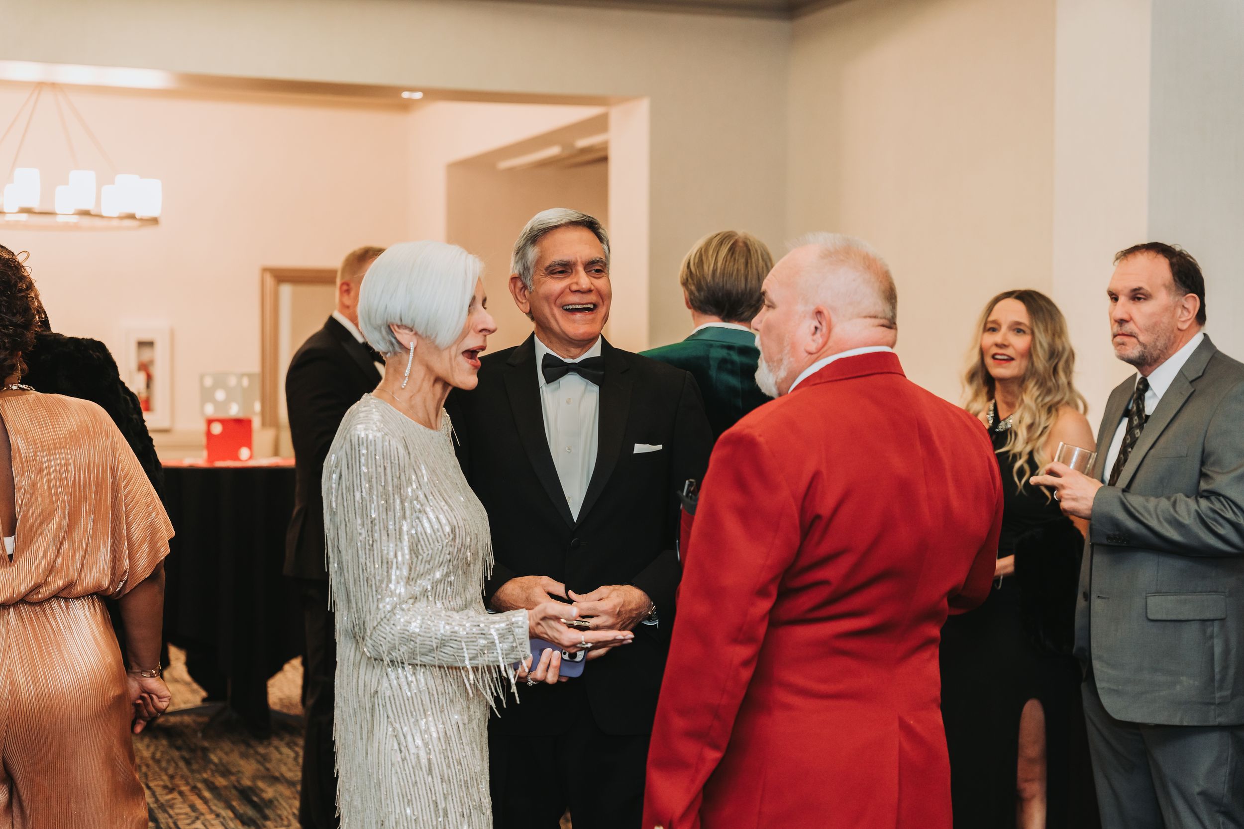 Elegantly dressed guests mingle at a formal gala event, featuring a woman in silver sequins and a man in a red tuxedo.