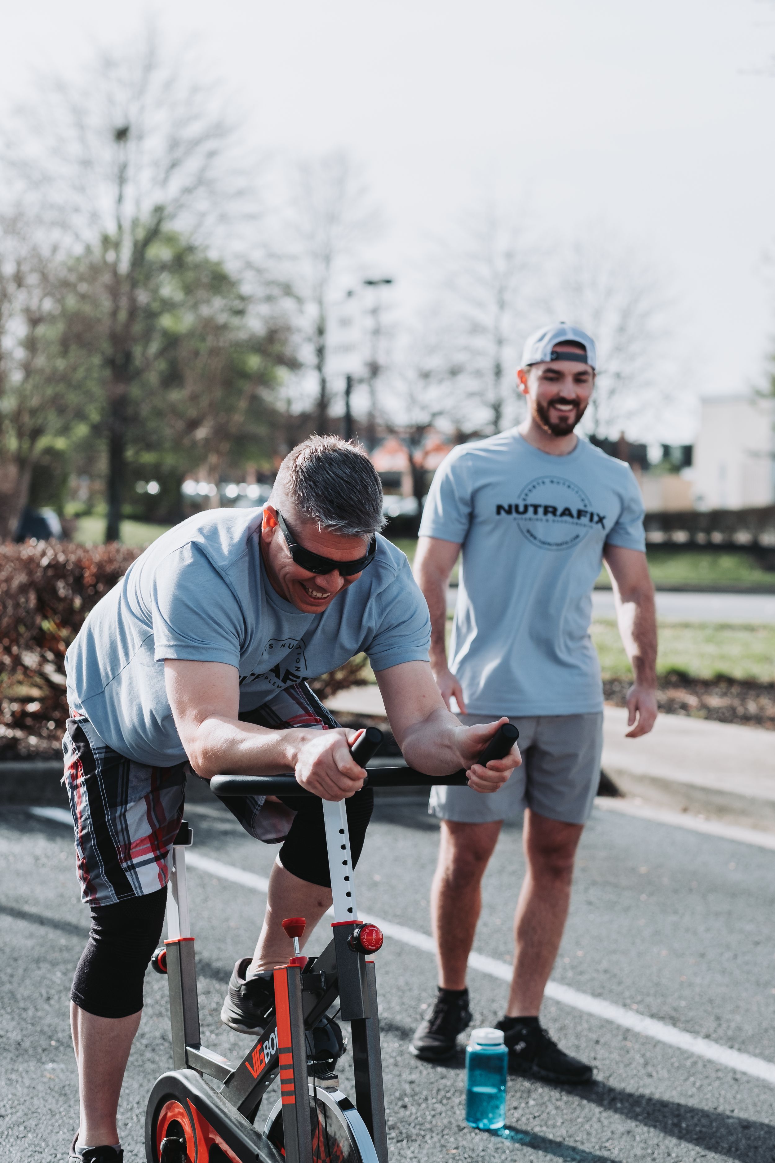 Two men in gray t-shirts exercise outdoors, one on a stationary bike, the other wearing a Nutrafix shirt smiling.