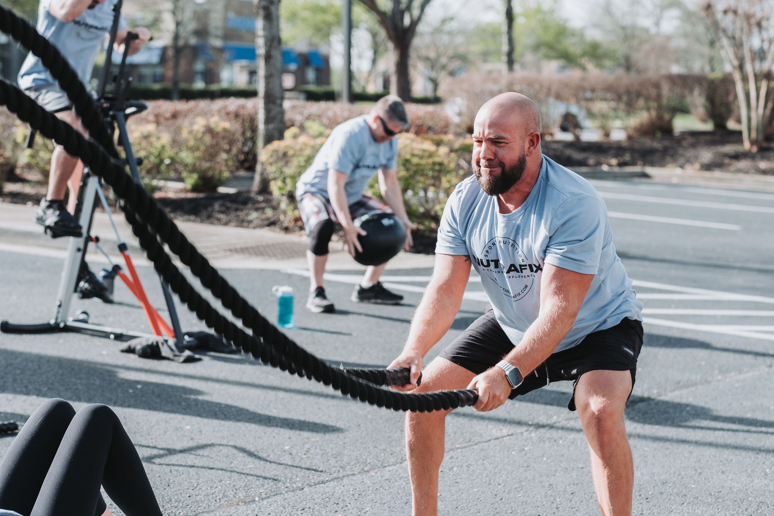 Man doing battle rope workout outdoors at fitness event with another athlete in background.