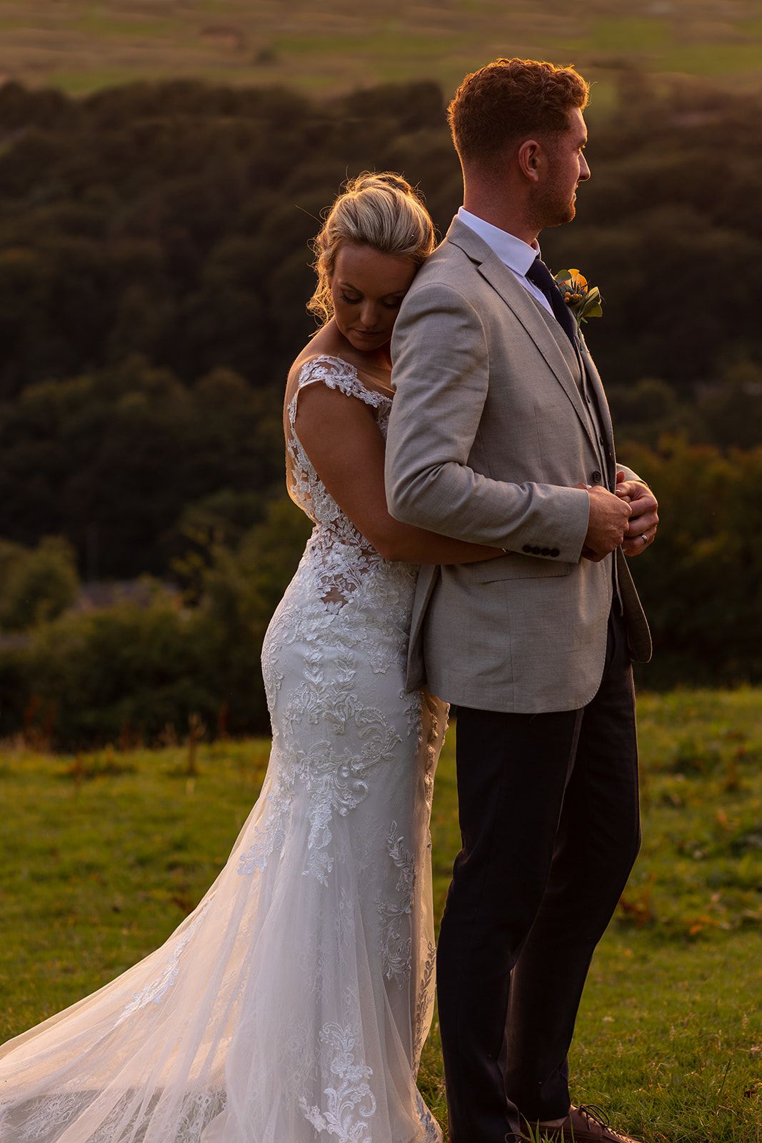 Bride in lace mermaid gown embraces groom in grey suit at golden hour with rolling green hills backdrop.