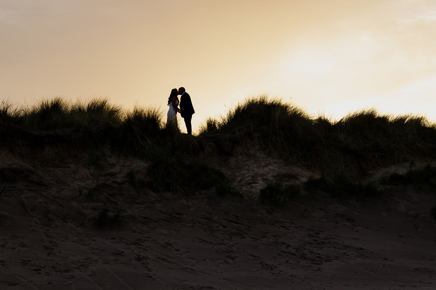 Silhouette of a couple kissing on sand dunes at sunset, with golden sky and coastal grasses in the background.