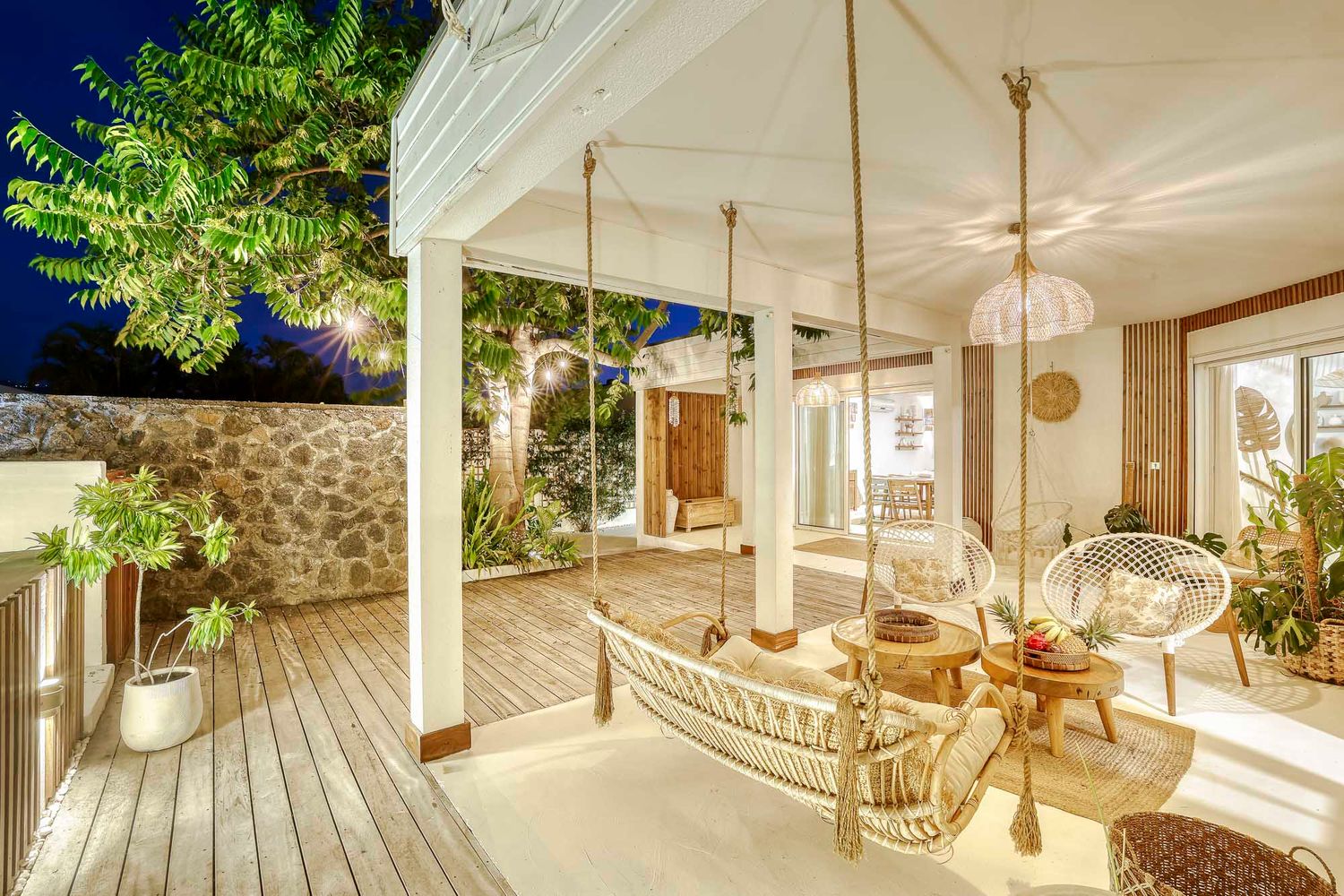 Boho outdoor terrace with hanging hammock chairs, wood deck, stone wall, and lush greenery at dusk.