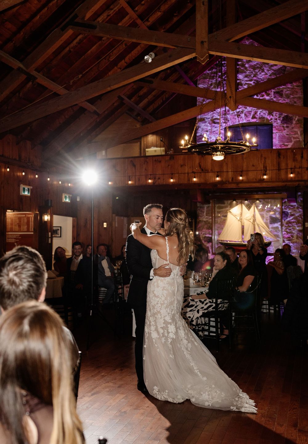 Bride and groom share their first dance in a rustic barn wedding venue with warm purple uplighting and exposed wooden beams.