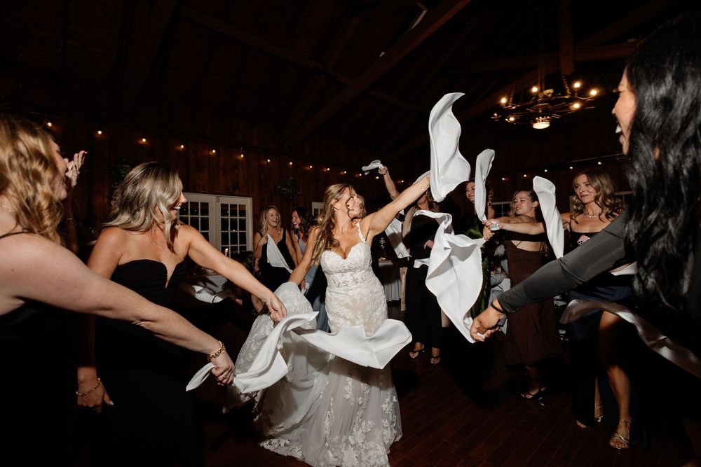 Bride dancing joyfully with bridesmaids waving white napkins at a wedding reception in a rustic venue.