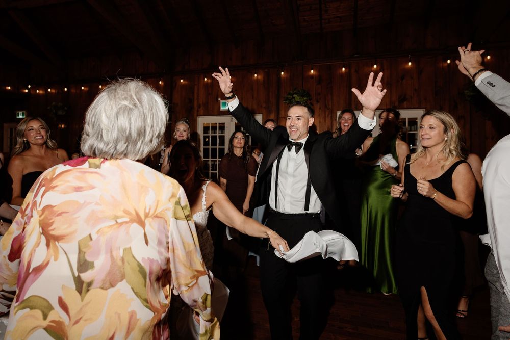 Groom in tuxedo dancing joyfully with guests at elegant wedding reception in rustic barn venue.