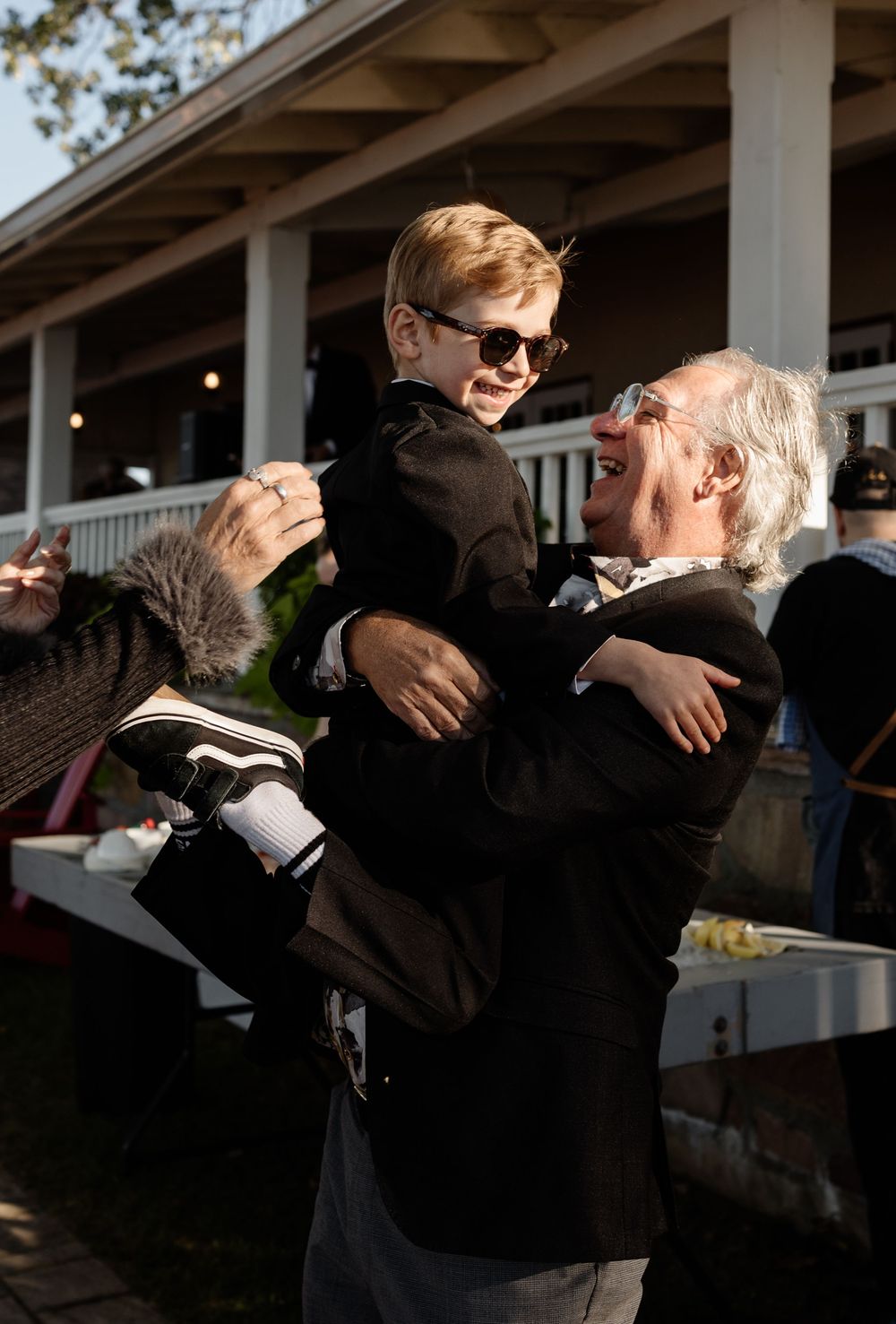 An elderly man joyfully lifts a young boy in a black suit wearing sunglasses outdoors at a formal event.