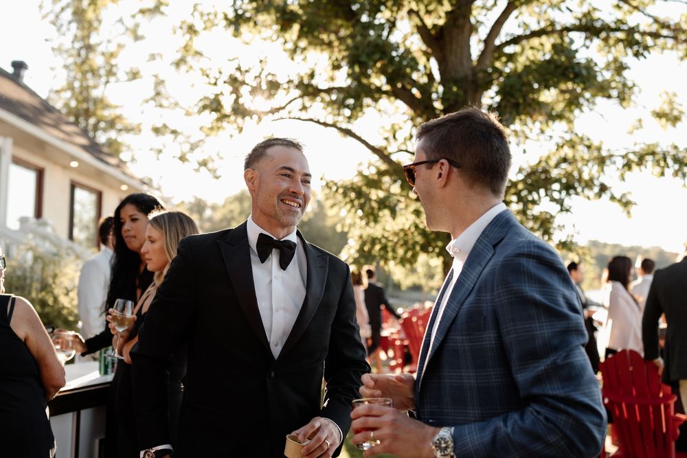 Two men in formal attire laughing at an outdoor wedding reception with guests and trees in background.