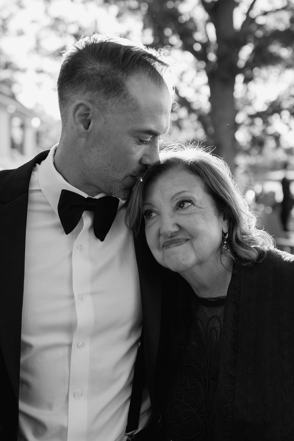 Black and white photo of a groom in a tuxedo sharing a tender moment with an older woman, likely his mother.