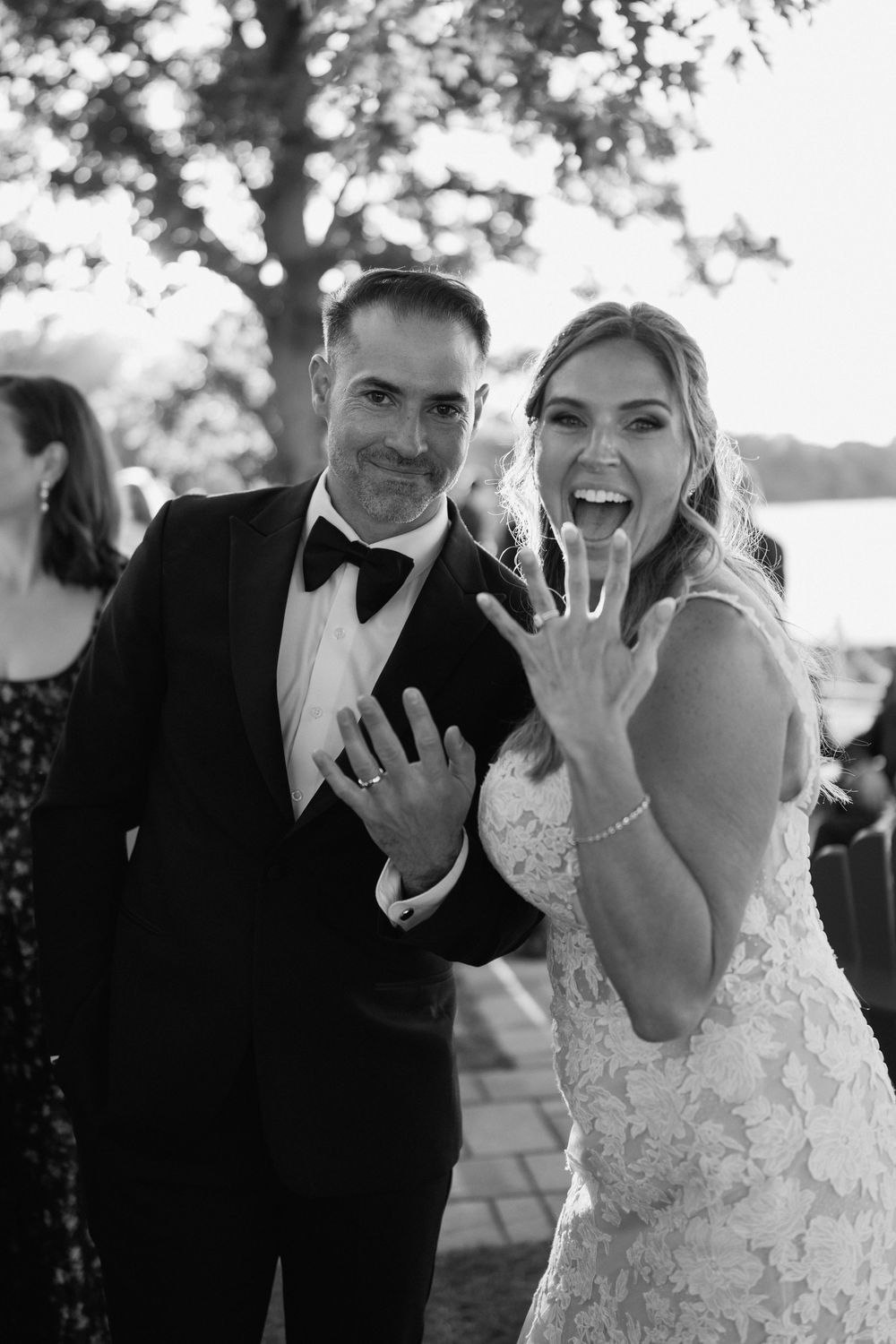 Black and white photo of newlyweds smiling and showing off wedding rings, bride in lace gown, groom in tuxedo.