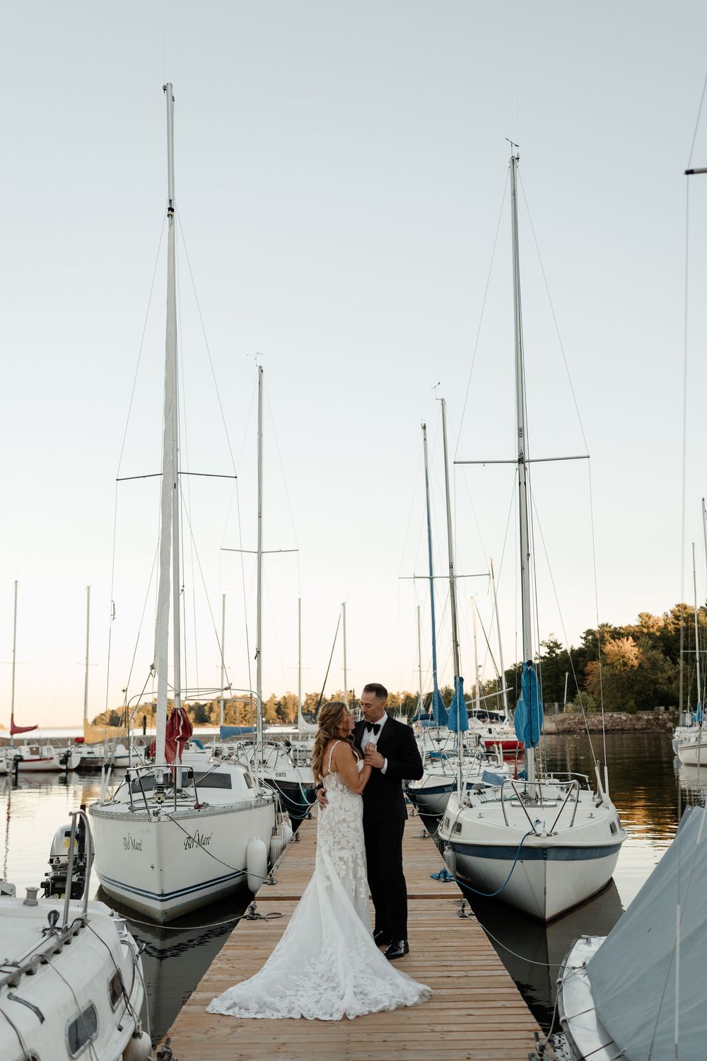 Bride and groom stand on a marina dock surrounded by sailboats at sunset in a romantic wedding portrait.