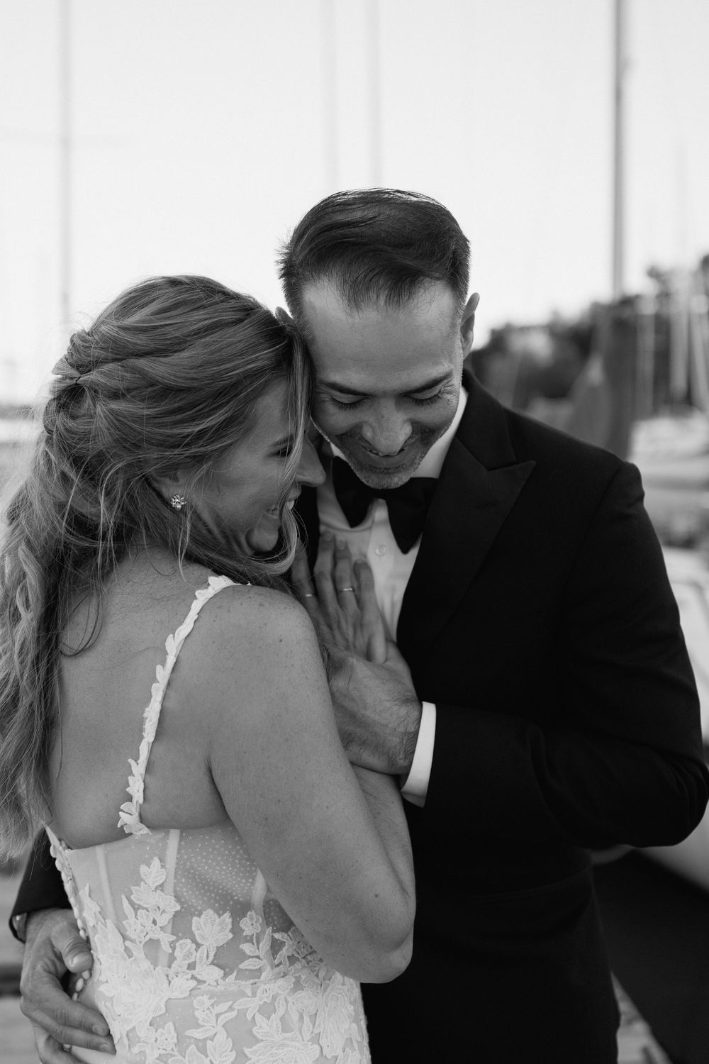 Black and white wedding photo of a laughing bride in lace gown embracing a smiling groom in a tuxedo.