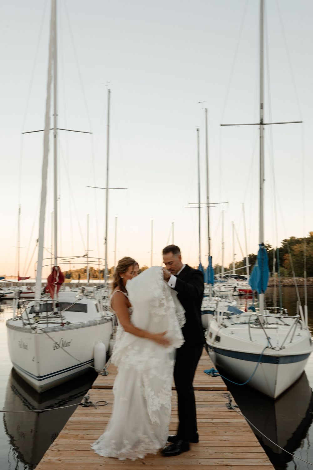 Bride and groom share a romantic moment on a marina dock surrounded by sailboats at golden hour sunset.