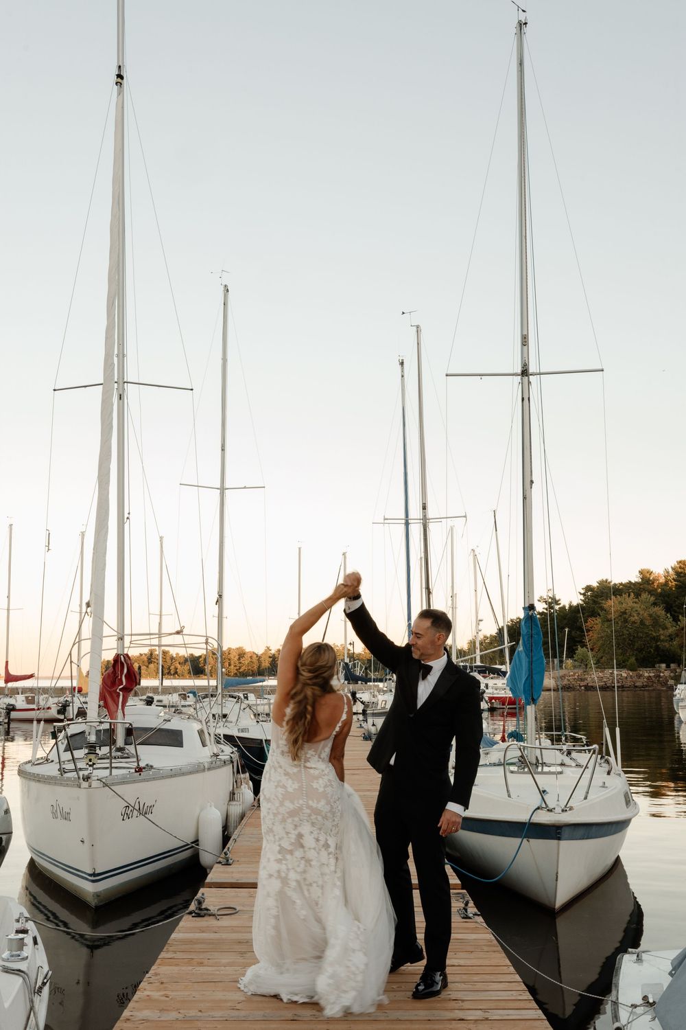 Bride and groom toast champagne on a wooden marina dock with sailboats at sunset on their wedding day.