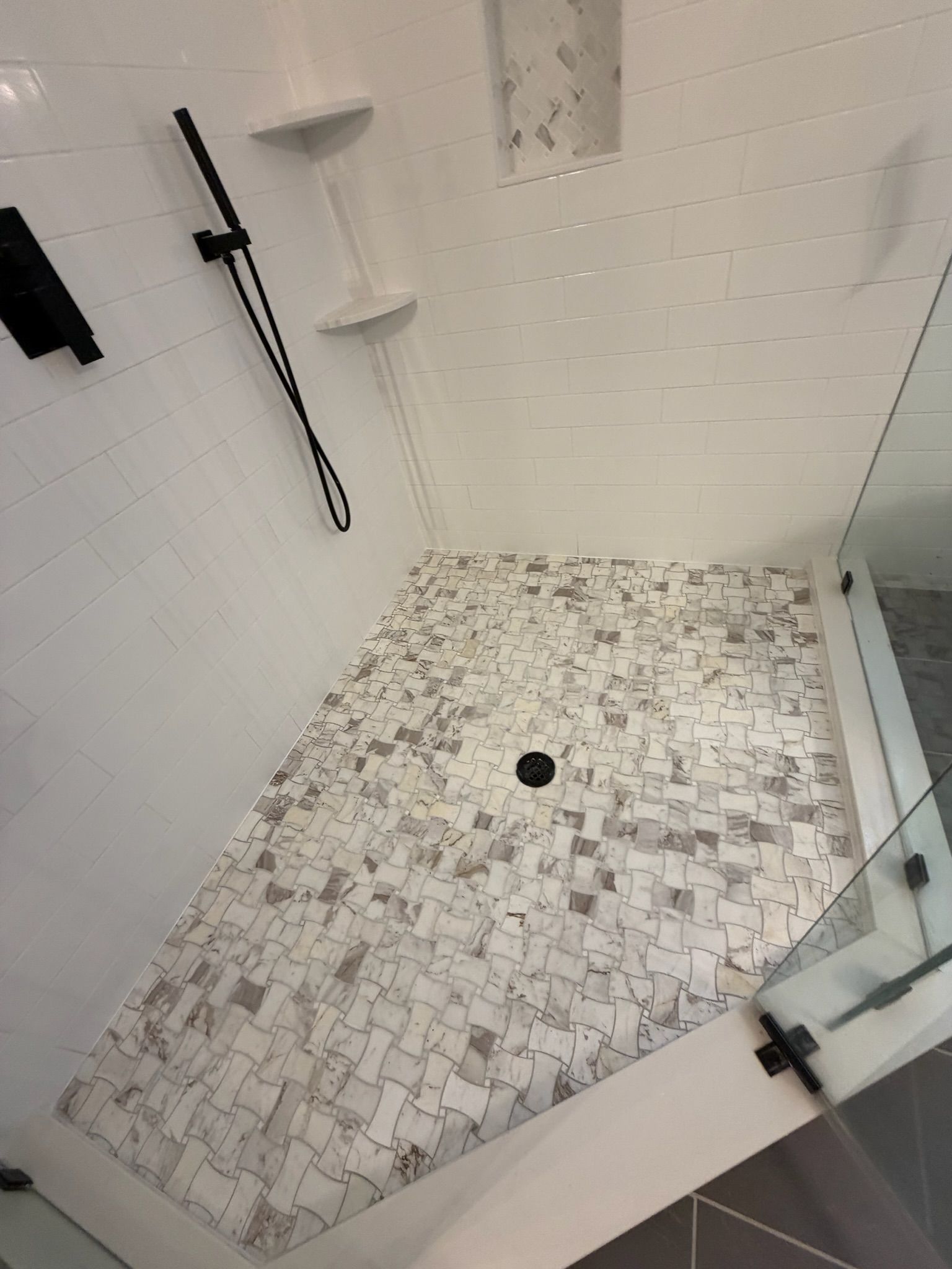 Corner shower with white subway tile walls and marble mosaic tile floor with black drain.