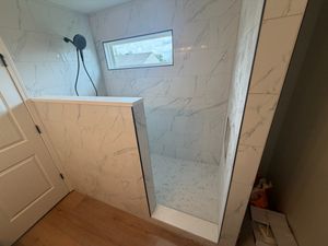 Walk-in shower with large white Calacatta marble-look porcelain tiles, half glass panel, and wood-look flooring nearby.