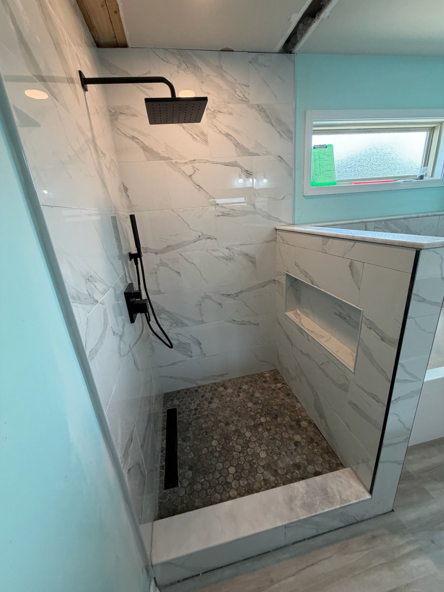 Walk-in shower with white marble porcelain tiles, matte black rain shower system, pebble floor, and glass panel enclosure.
