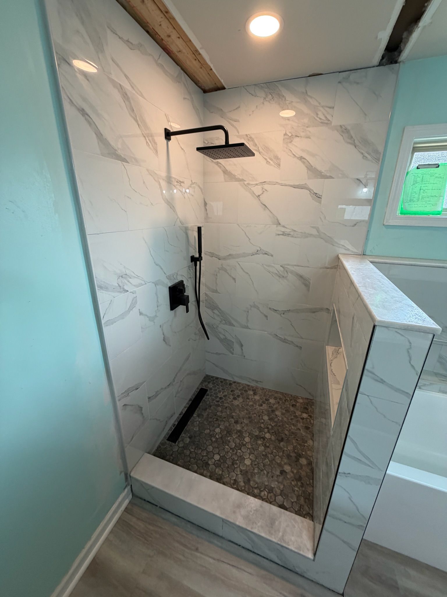 Modern walk-in shower with Calacatta marble tiles, matte black rain showerhead, pebble tile floor, and exposed wood beam accent.