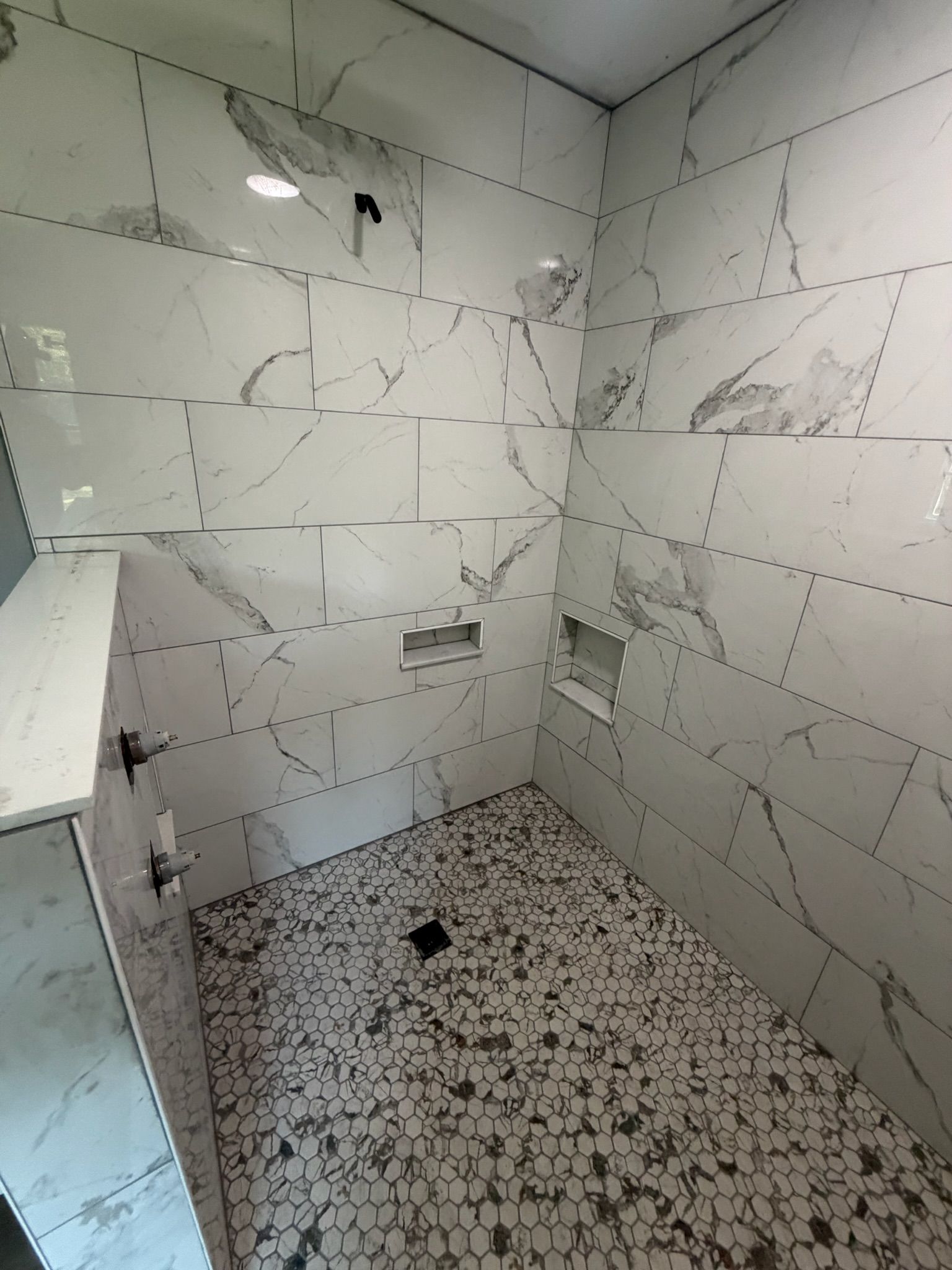 Luxury walk-in shower with large white marble-look porcelain wall tiles and hexagonal mosaic floor tiles with black drain.