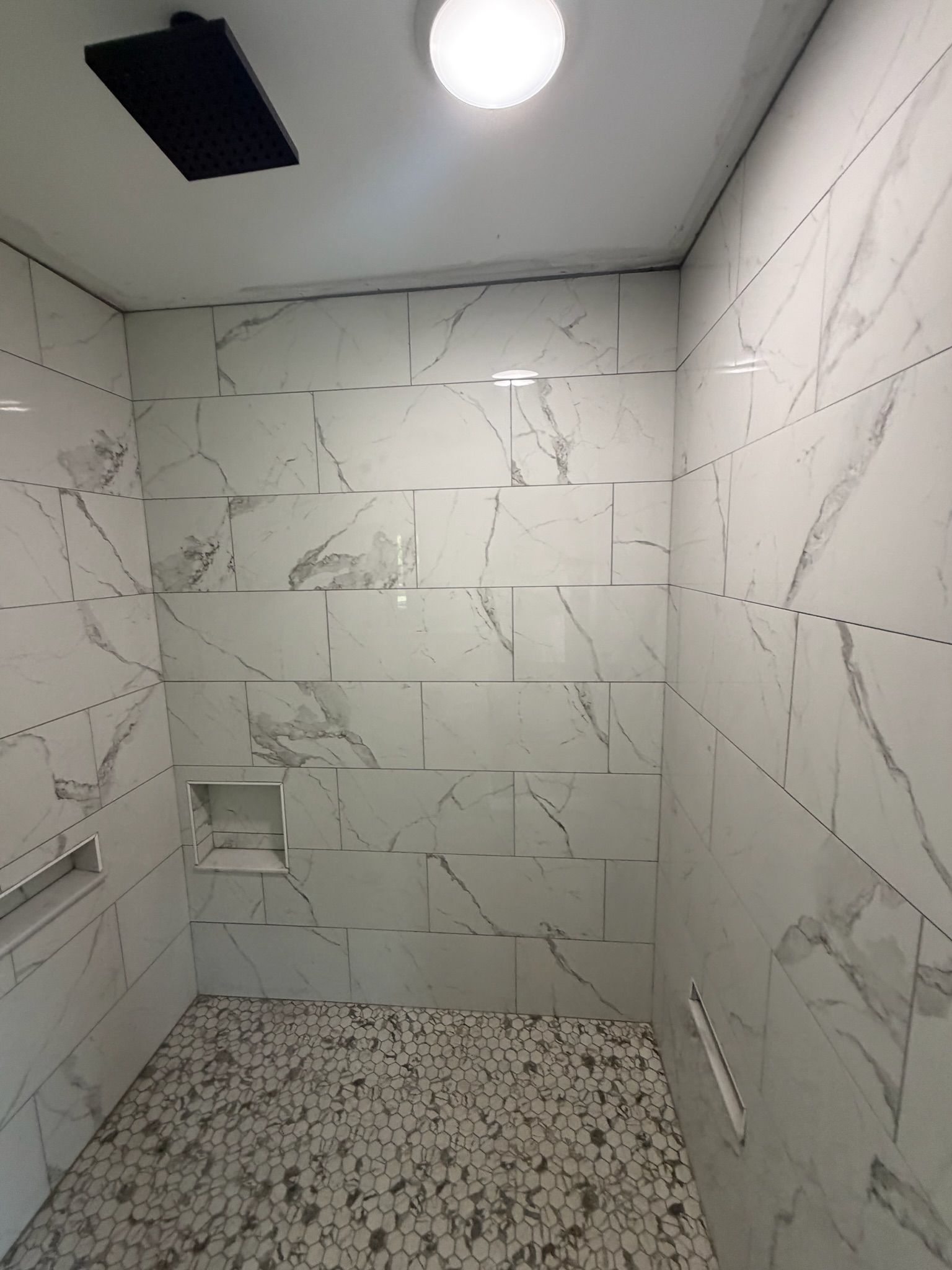 Walk-in shower with large white marble-look porcelain wall tiles, built-in niche shelves, and white pebble stone floor.