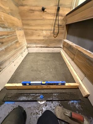 Worker leveling a concrete shower floor with a spirit level, surrounded by wooden walls during bathroom renovation construction.