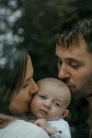 Mother and father kissing newborn baby behind window in Corbridge