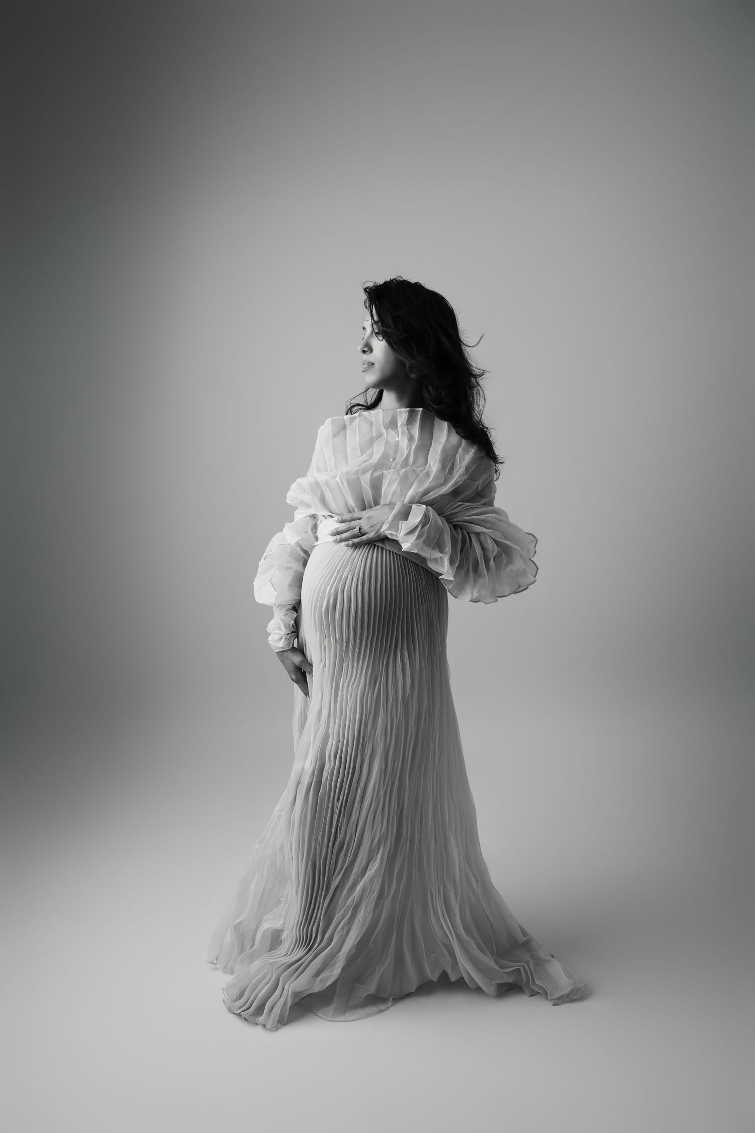 Black and white maternity photo of a brunette woman in elegant pleated gown holding her baby bump in a studio.