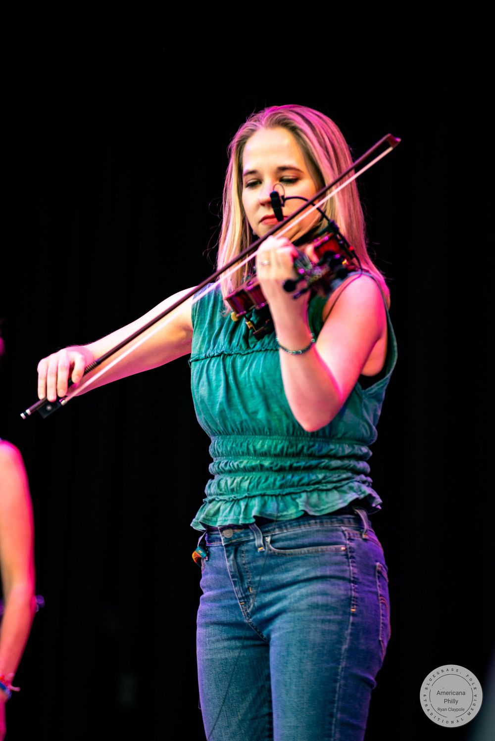 Young woman playing violin on stage, wearing a green sleeveless top and jeans under colorful performance lighting.