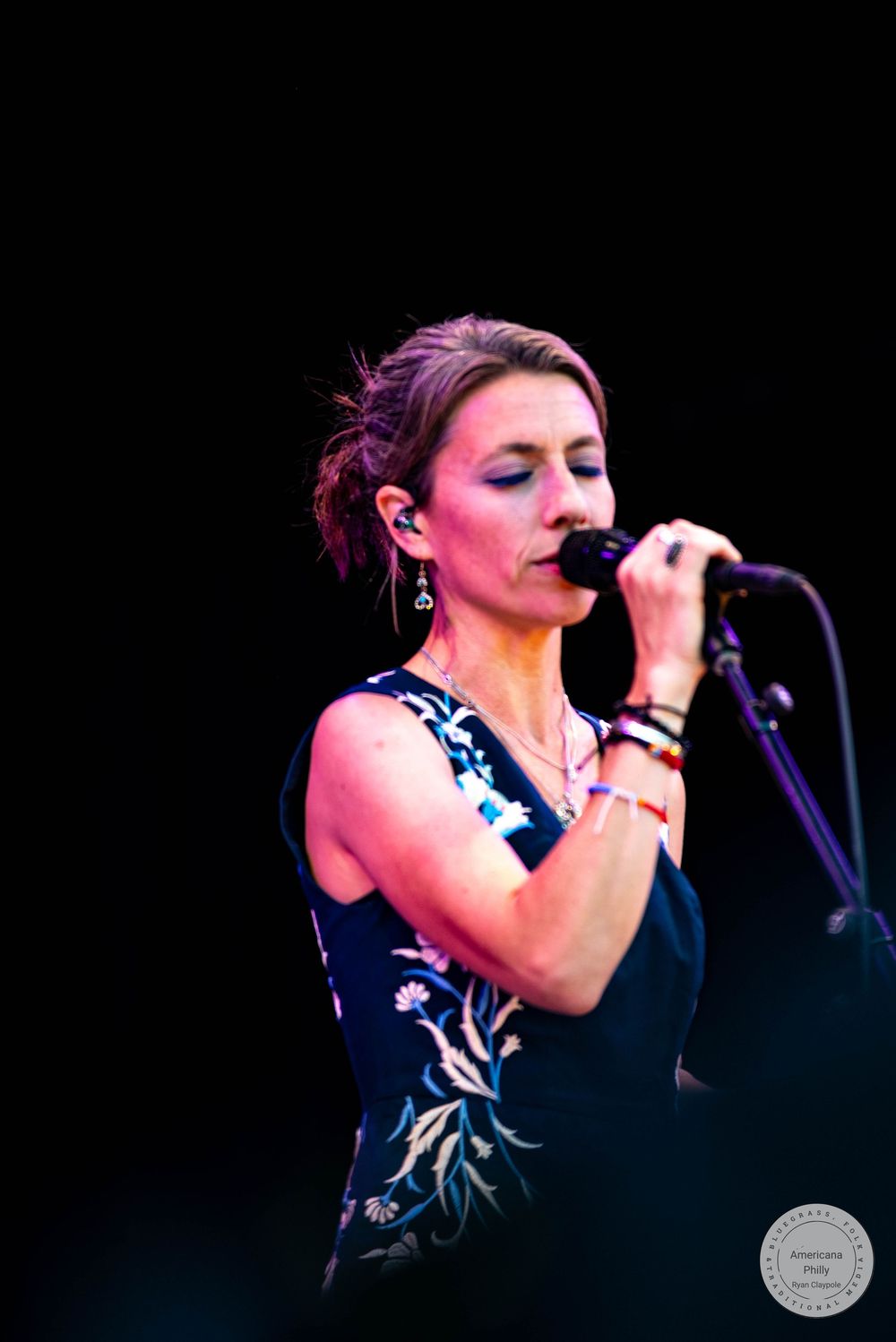 Woman singer performing on stage, holding microphone, wearing floral dress and earrings, eyes closed, concert setting.