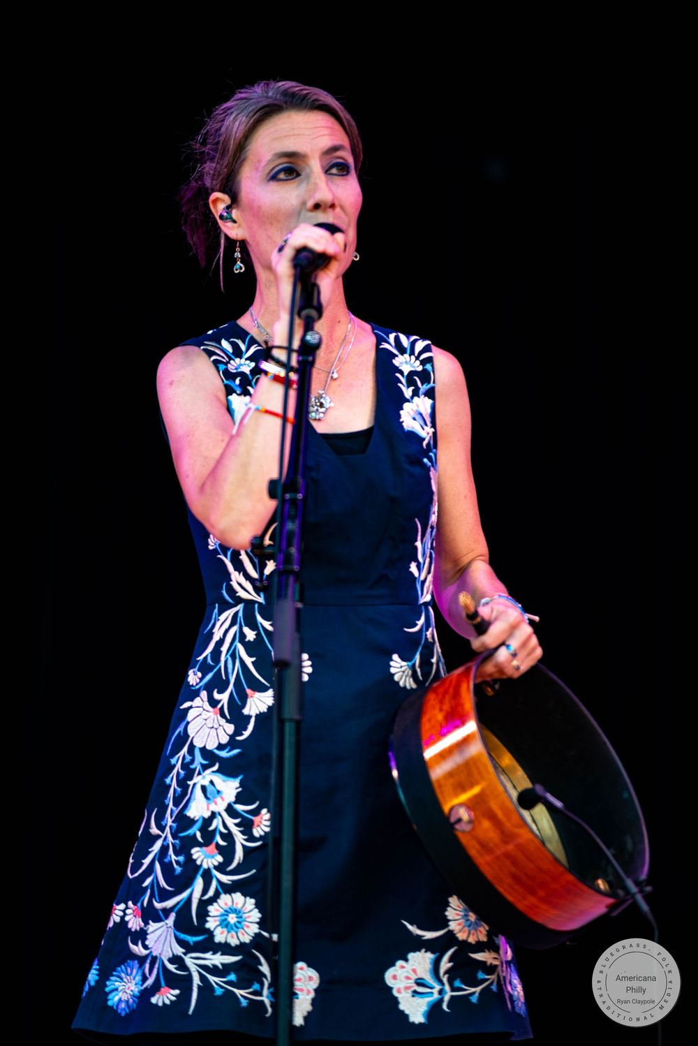 Woman in floral dress singing at microphone while holding a tambourine on stage during a live performance.