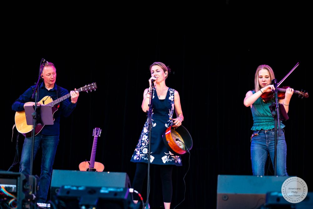 Three women performing on stage, one playing guitar, one singing with tambourine, and one playing violin.
