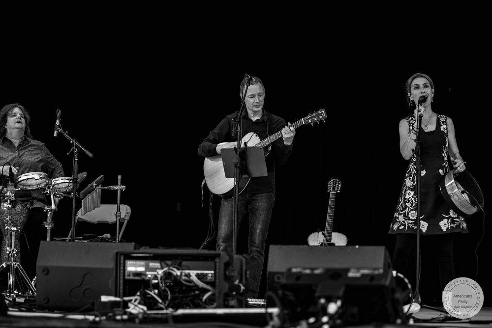 Three women performing live on stage, one playing acoustic guitar, one singing, and one on drums, in black and white.