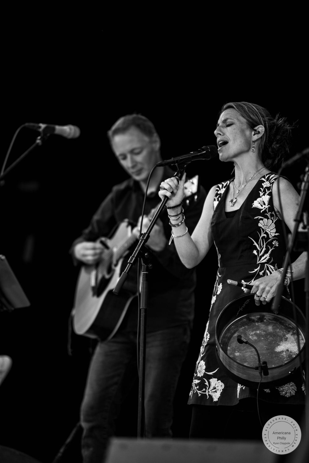 Black and white photo of a woman singing into a microphone holding a tambourine with a guitarist performing behind her.
